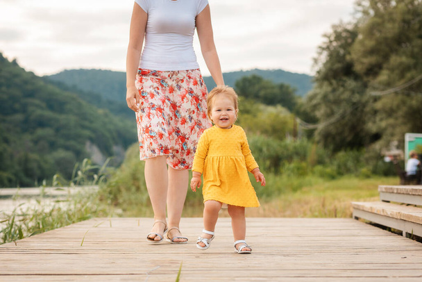 Feliz niña caucásica en vestido amarillo caminando por el muelle del río con su madre. Al aire libre. Concepto de día del niño. - Foto, imagen