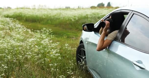 Mujer fotógrafa sentada en el coche y fotografiando un paisaje de campo de flores - Imágenes, Vídeo