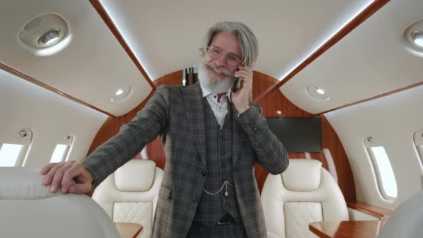 Portrait d'un homme millionnaire senior élégant parlant sur un téléphone portable, debout dans un avion privé. Homme d'affaires PDG barbu souriant ayant un appel téléphonique dans un jet privé de luxe. - Séquence, vidéo
