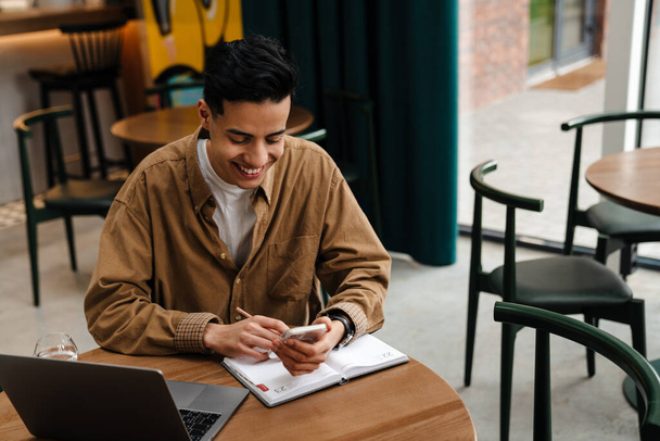 Young smiling hispanic man student sitting at the cafe table with laptop computer indoors, holding mobile phone - Fotografie, Obrázek