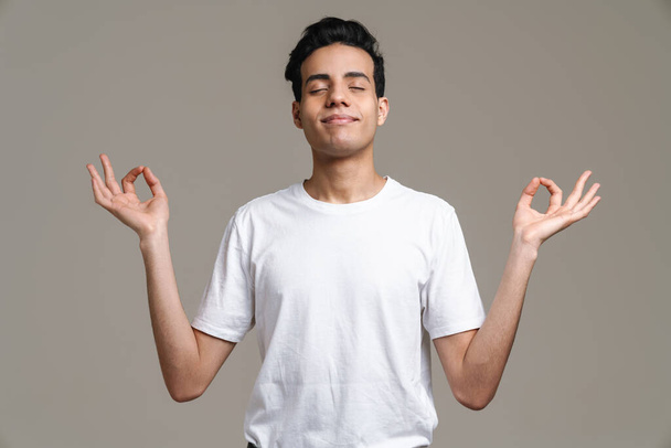 Usmívající se hispánec v tričku meditující a gestikulující izolovaně nad šedým pozadím - Fotografie, Obrázek