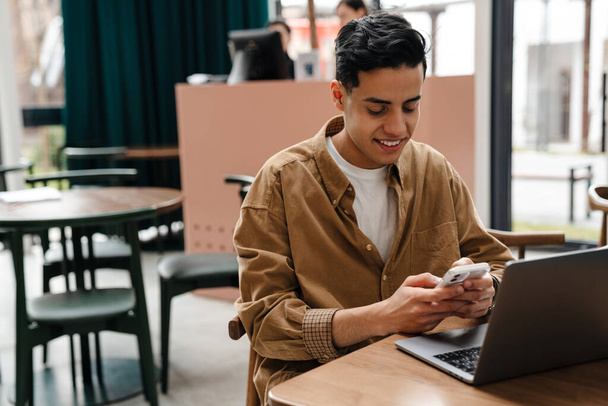 Young smiling hispanic man student sitting at the cafe table with laptop computer indoors, holding mobile phone - Photo, image