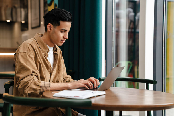 Jeune homme calme hispanique étudiant assis à la table de café avec ordinateur portable à l'intérieur - Photo, image