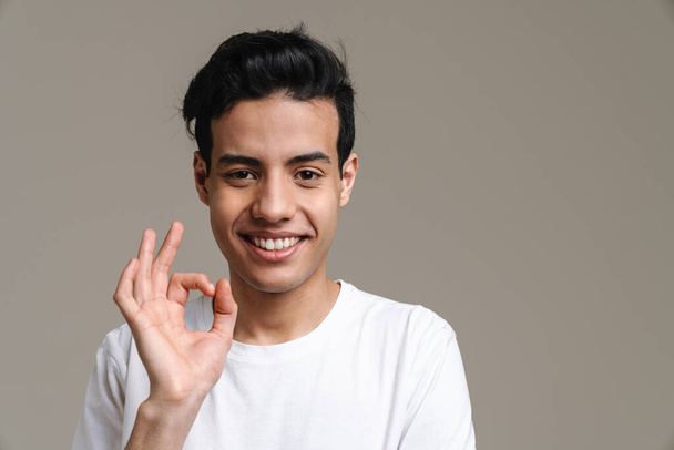 Brunette hispanic man in t-shirt smiling and gesturing at camera isolated over grey background - Photo, image