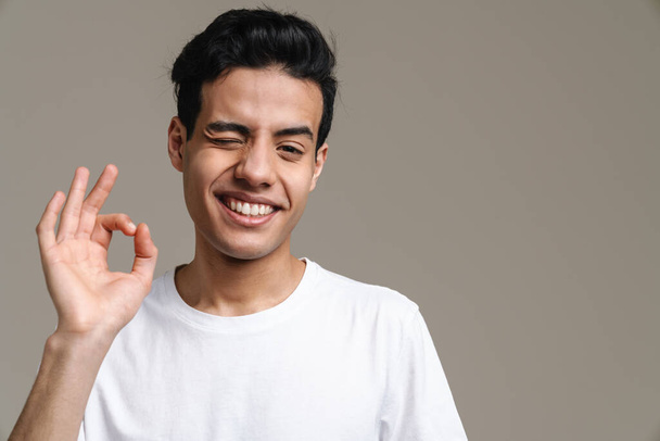 Brunette hispanic man in t-shirt winking and gesturing at camera isolated over grey background - Fotoğraf, Görsel