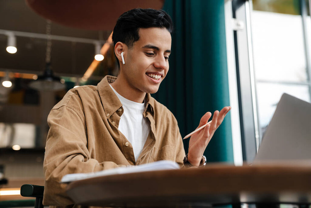 Jonge glimlachende Spaanse man student zittend aan de cafe tafel met laptop computer binnen, op een video gesprek - Foto, afbeelding