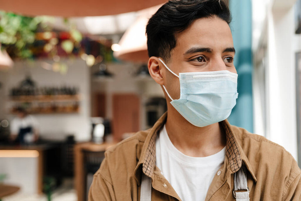 Koruyucu tıbbi maskeli ve önlüklü İspanyol genç bir garsonun kapalı bir kafede durduğu yeri kapatın. - Fotoğraf, Görsel