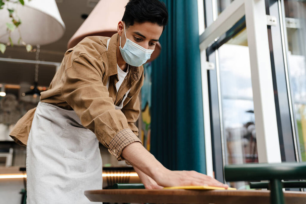 Young hispanic waiter disinfecting table in cafe while wearing face mask - Φωτογραφία, εικόνα