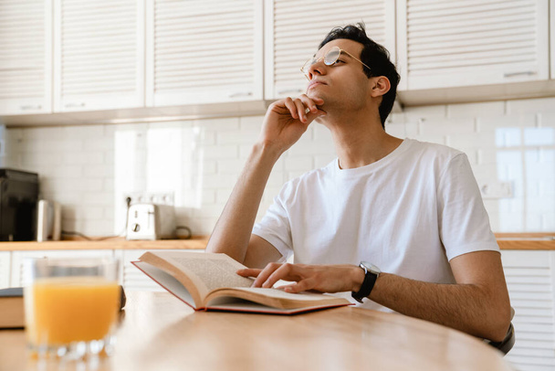 Young hispanic man reading a book while having breakfast in the kitchen at home, sitting at the table - Photo, Image