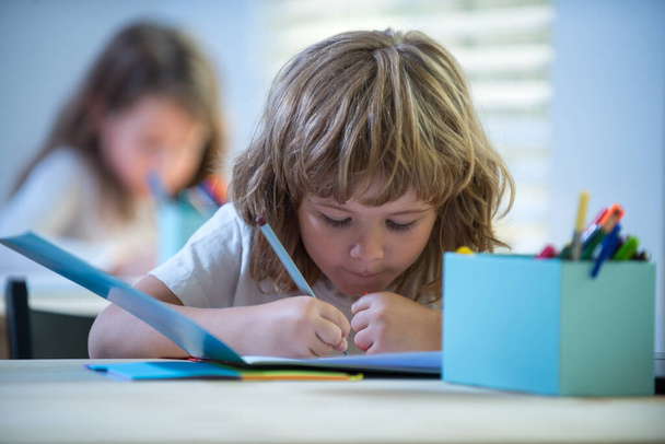 School kid writing something in copybook and sitting at table in classroom. Back to school. Happy smiling pupil drawing at the desk. Child from primary school in the classroom. - Zdjęcie, obraz