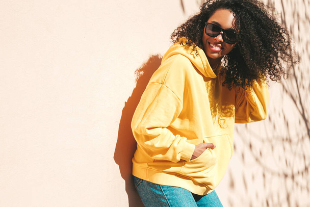 Hermosa mujer negra con peinado afro rizos. Modelo hipster sonriente en sudadera con capucha amarilla. Sexy mujer despreocupada posando en el fondo de la calle en gafas de sol. Alegre y feliz - Foto, Imagen