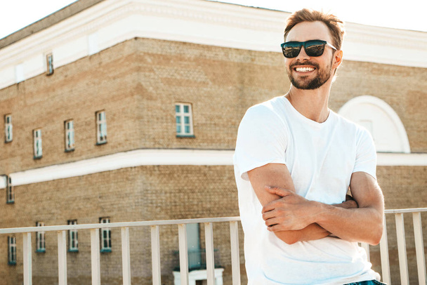 Portrait of handsome smiling  hipster lambersexual model.Stylish man dressed in white T-shirt and jeans. Fashion male posing on the street background near fence at the beach  - Foto, immagini