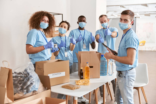 Group of diverse people wearing blue uniform, protective masks and gloves showing thumbs up while sorting donated food items, volunteering in community together - Photo, Image