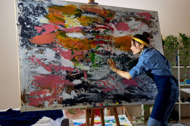 Creative young female painter working on a large modern abstract oil painting, applying paint on canvas with fingers in living room at home studio workshop - Foto, Bild