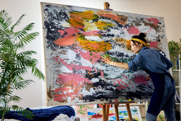 Inspirált női művész dolgozik egy nagy modern absztrakt olajfestmény, alkalmazó festék vászonra ujjakkal, áll otthon stúdió műhely - Fotó, kép