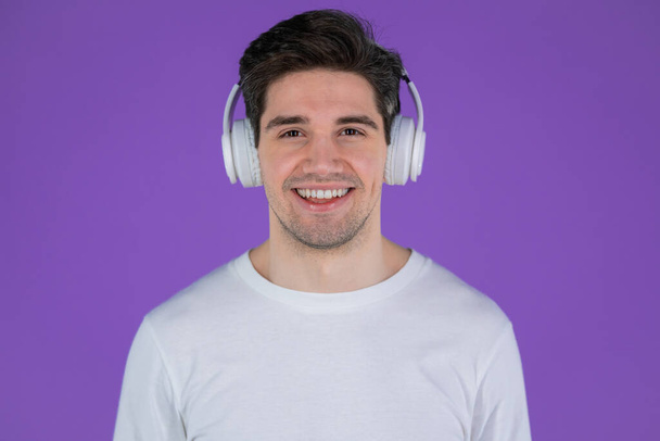 Charming young man listening to music with wireless headphones, guy having fun, smiling in studio on purple background. Dance, radio concept. - Valokuva, kuva