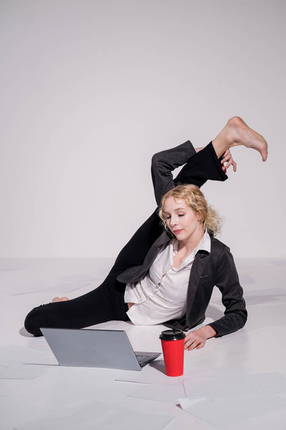 Barefoot ballerina dressed in a business suit poses for a laptop and drinks coffee. Flexible woman works at the computer - Фото, изображение