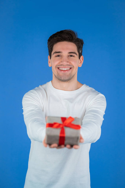 Handsome man holding gift and gives it by hands to camera. He is happy, smiling. Guy on blue background. Positive holiday photo - Foto, Imagem