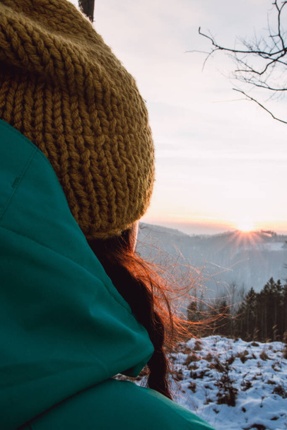 Hermosa joven de 20-25 años mirando una hermosa puesta de sol de invierno en las montañas de la República Checa. Fin del día. Gorra amarilla y chaqueta verde. - Foto, Imagen