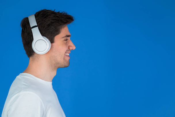 Young man listening to music with wireless headphones, guy having fun, smiling in studio on blue background. Copy space. Dance, radio concept. - Foto, Bild