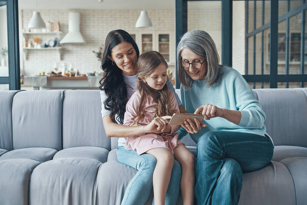 Little girl with mother and grandmother using smart phone while sitting on the sofa at home - Foto, immagini