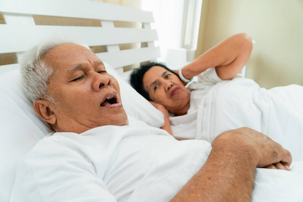 Ältere Asiatin, die ihre Augen und Ohren offen legt und mit den Händen im Bett neben ihrem Mann liegt, der schnarcht und Lärm macht, Eheprobleme. - Foto, Bild