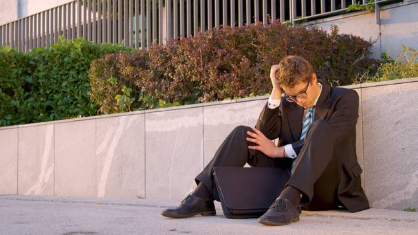 Depressed young Caucasian yuppie sitting on the sidewalk after going bankrupt. - Foto, Imagen