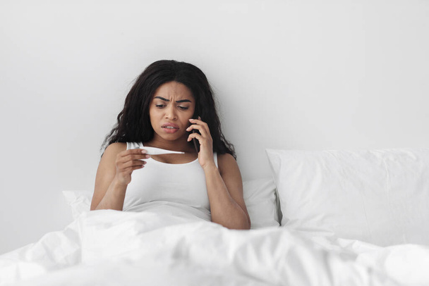 Aggódó afro-amerikai nő ül az ágyban, kezében a hőmérő és hívja a háziorvos tanácsadás - Fotó, kép