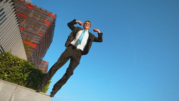 Nízký úhel: Usmívající se bělošský podnikatel skočí z římsy s nataženýma rukama. - Fotografie, Obrázek