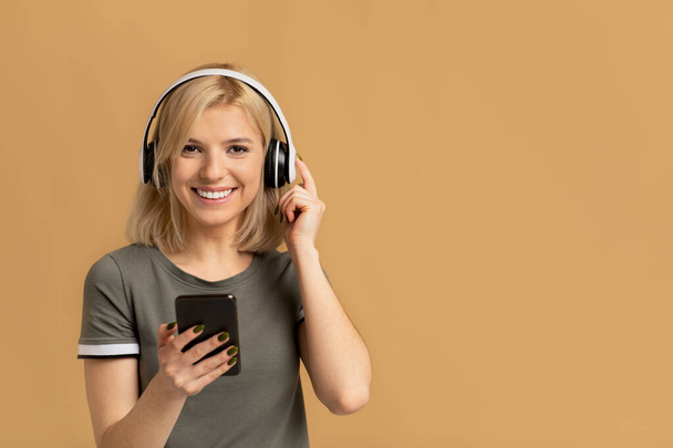 Portrait of emotional lady listening to music in wireless headset, standing isolated on peach studio background - Foto, imagen