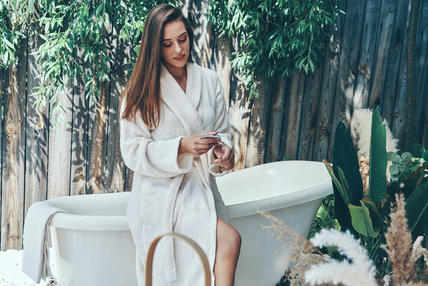 Beautiful young woman moisturizing hand cream while leaning at the bathtub outdoors - Фото, изображение