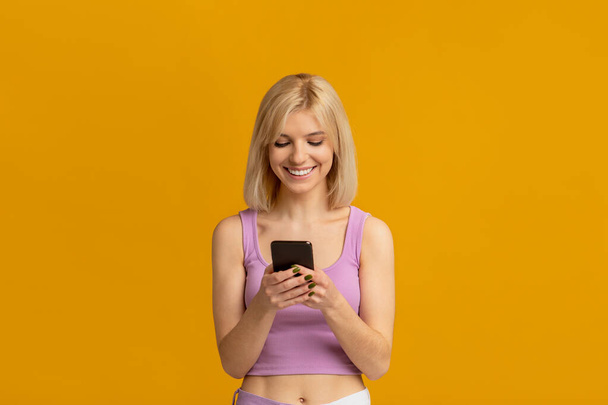 Rozmawiam z przyjaciółmi. Szczęśliwa młoda kobieta stojąca ze smartfonem w dłoni nad żółtym tle studio - Zdjęcie, obraz