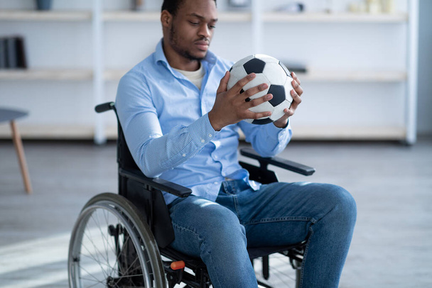 Hombre negro discapacitado con pelota de fútbol sentado en silla de ruedas, sintiéndose deprimido por su lesión en casa, enfoque selectivo - Foto, imagen