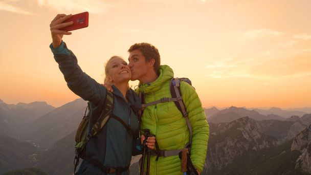 CLOSE UP: Goofy young man kisses his girlfriend on the cheek taking a selfie. - Foto, Bild