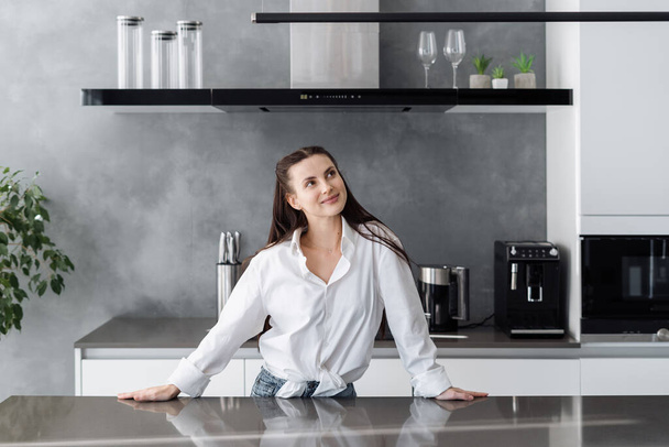 Young beautiful dreamy woman in casual outfit standing at table in modern fully furnished kitchen and thinking what to cook on breakfast for whole family on weekend - Foto, immagini