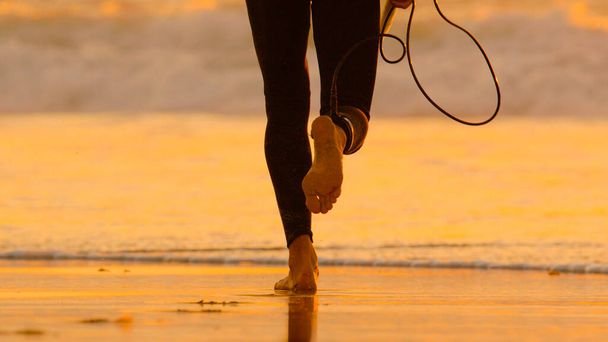 LOW ANGLE: Unerkennbarer Surfer im Neoprenanzug rennt dem schäumenden Ozean entgegen. - Foto, Bild