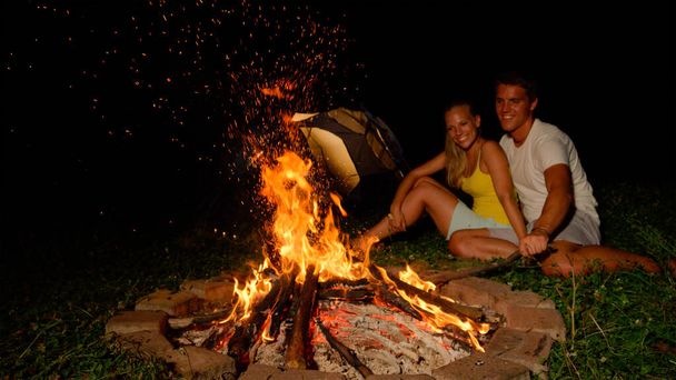 CLOSE UP: Cheerful man strikes campfire with stick while snuggling with girl. - Фото, изображение