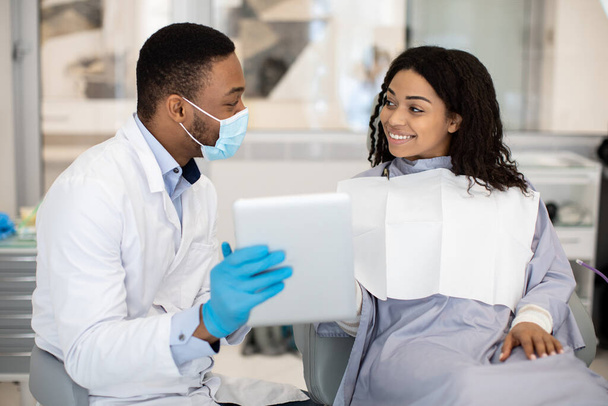 Zwarte mannelijke tandarts toont tandenfoto op digitale tablet aan vrouwelijke patiënt - Foto, afbeelding