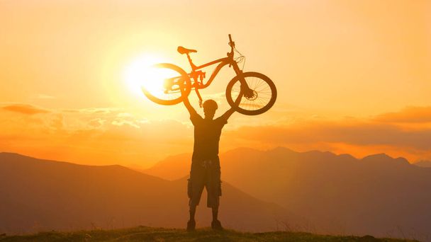 Nuori miespyöräilijä pitää pyöränsä yläpuolella kultaisella auringonlaskulla.. - Valokuva, kuva