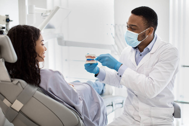 Schwarzer Stomatologe hält Plastikkiefer und lehrt Patientin Dentalhygiene - Foto, Bild