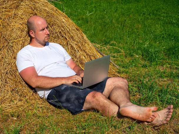 Tişört ve şort giymiş orta yaşlı bir adam elinde dizüstü bilgisayarla sarı bir samanlıkta yatıyor. Uzaktan çalışma, eğitim. Kırsal alan - Fotoğraf, Görsel