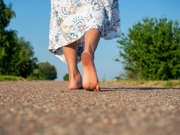 Een vrouw gekleed in een jurk loopt blootsvoets over het asfalt. Achteraanzicht. Het concept van reizen, recreatie, vakantie, vrijheid - Foto, afbeelding