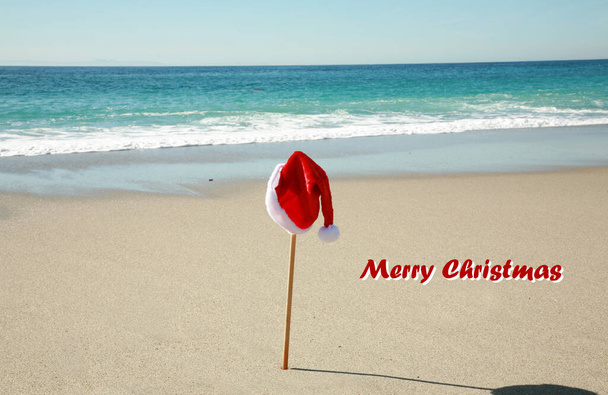 Chapéu Papai Noel. Um chapéu de Pai Natal num poste na praia. Papai Noel tira o chapéu quando vai nadar no oceano. Feliz Natal para todos. Boas festas. Papai Noel chapéu de pele vermelha pendura em um pólo na praia. Ho Ho Ho.  - Foto, Imagem