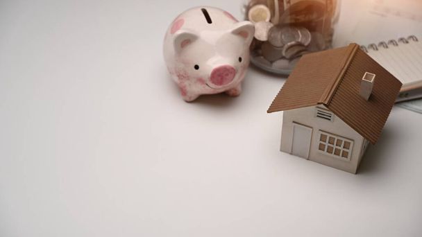 Sparschwein, Münze und Hausmodell auf weißem Tisch. Sparen für die Zukunft, Sparen für den Haus- oder Immobilienmarkt. - Foto, Bild