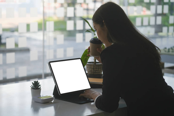 Οπίσθια όψη της γυναίκας επιχειρηματία με κοστούμι πίνοντας ζεστό καφέ και την εργασία με tablet υπολογιστή στο γραφείο. - Φωτογραφία, εικόνα
