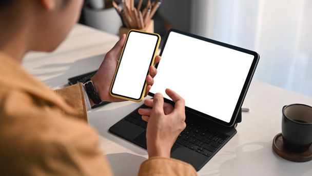 Plan recadré d'un jeune homme lisant un courriel sur une tablette d'ordinateur et utilisant un téléphone mobile au bureau à domicile. - Photo, image