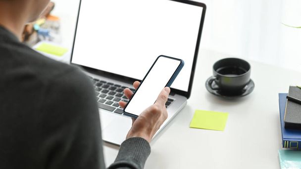 Olkapään yli näkymä nuoren miehen freelancer matkapuhelimella ja työskentelee tietokoneen kannettavan nykyaikaisella työpaikalla. - Valokuva, kuva