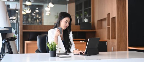 Горизонтальный образ деловой женщины, пьющей кофе и работающей с планшетным компьютером в офисе. - Фото, изображение
