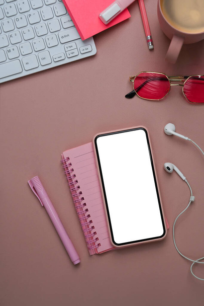 Nad pohledem na ženský pracovní prostor s chytrým telefonem, sluchátkem, notebookem a klávesnicí na pastelově růžovém pozadí. - Fotografie, Obrázek