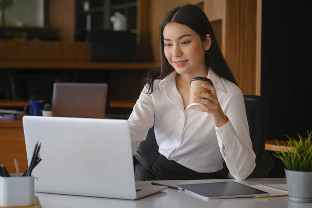 Lächelnde Geschäftsfrau hält Kaffeetasse in der Hand und arbeitet am Laptop. - Foto, Bild